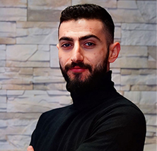 Ein junger Mann mit dunklem Vollbart und schwarzem Rollkragenpullover lächelt selbstbewusst vor einer Wand aus hellen Steinen