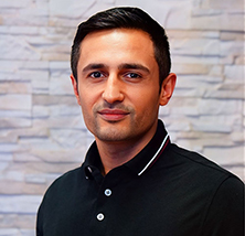 Ein Mann mit kurzen dunklen Haaren und ernstem Blick trägt ein schwarzes Poloshirt vor einer hellen Steinwand