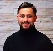 Ein lächelnder Mann mit Bart und dunklen Haaren trägt einen schwarzen Rollkragenpullover vor einer Wand aus hellen Steinen