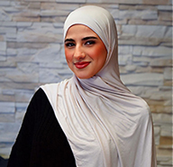 Junge Frau mit hellem Kopftuch lächelt freundlich vor einer grauen Steinwand