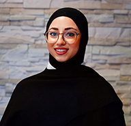 Junge Frau mit schwarzem Kopftuch und Brille lächelt vor heller Steinwand