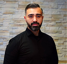 Ein Mann mit dunklem Bart und ernster Miene vor einer hellen Steinwand im schwarzen Hemd