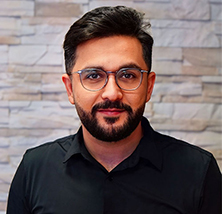 Ein Mann mit Brille und Bart lächelt leicht vor einer Wand mit Steinmuster