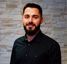 Mann mit dunklem Bart und schwarzem Hemd lächelt leicht vor heller Steinwand